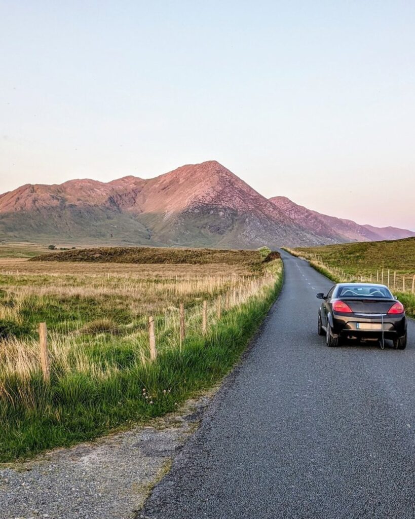 Black car driving on narrow road through rural Irish moorland with mountain backdrop