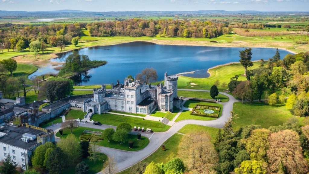 Aerial view of a large stone castle beside a blue lake surrounded by green fields and trees