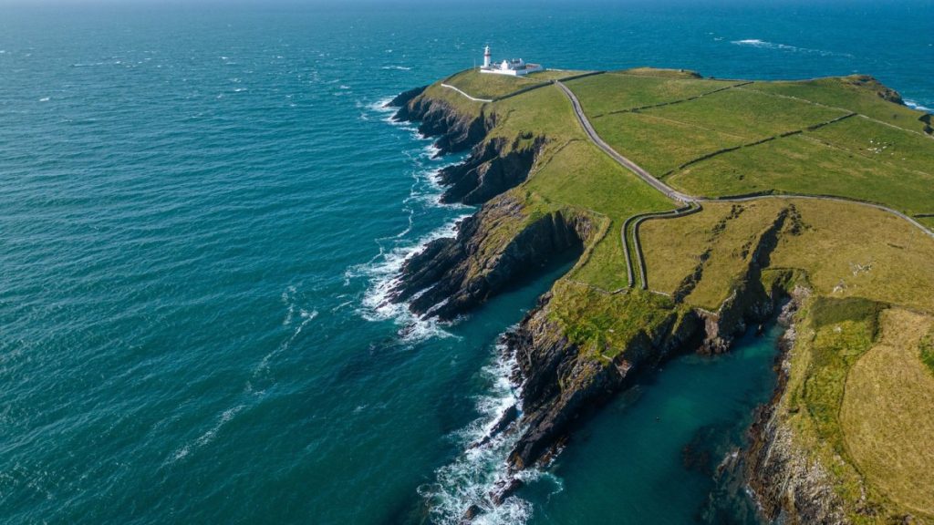 Aerial view of dramatic green coastal cliffs with winding road and lighthouse
