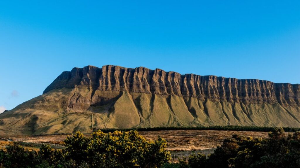 Dramatic striated mountain face rising above golden moorland under clear blue sky