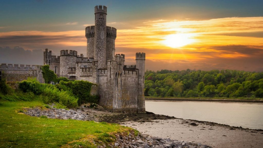 Medieval stone castle with towers on grassy riverbank at golden sunset